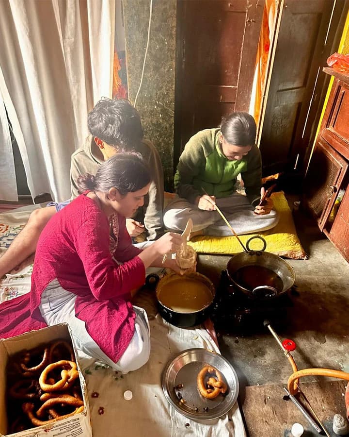 People sitting on the floor preparing Sel Roti batter and setting up a frying pot indoors with raw Sel Rotis in a box